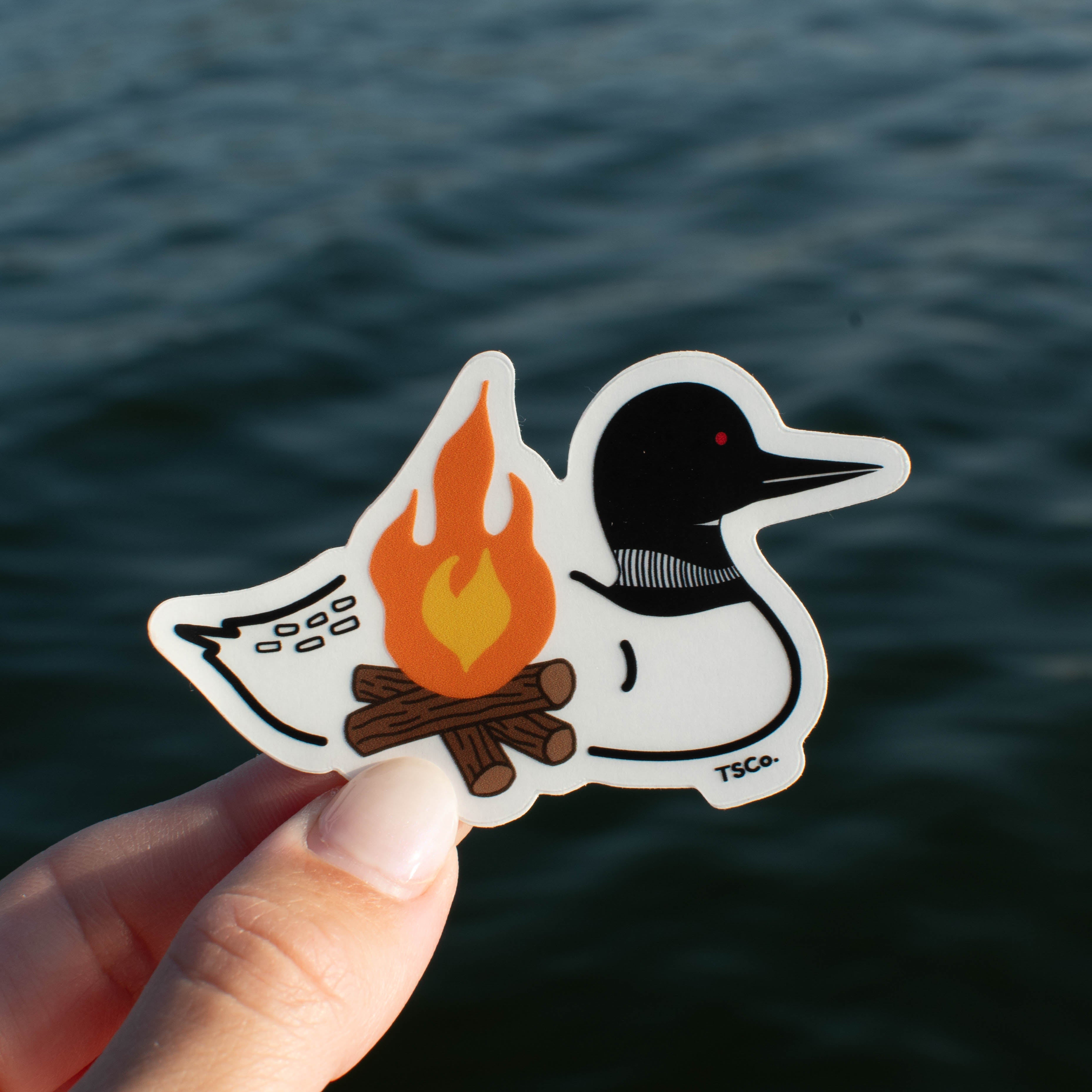 A hand holds a die-cut sticker featuring a stylized loon with a campfire, set against a blurred background of dark blue water.