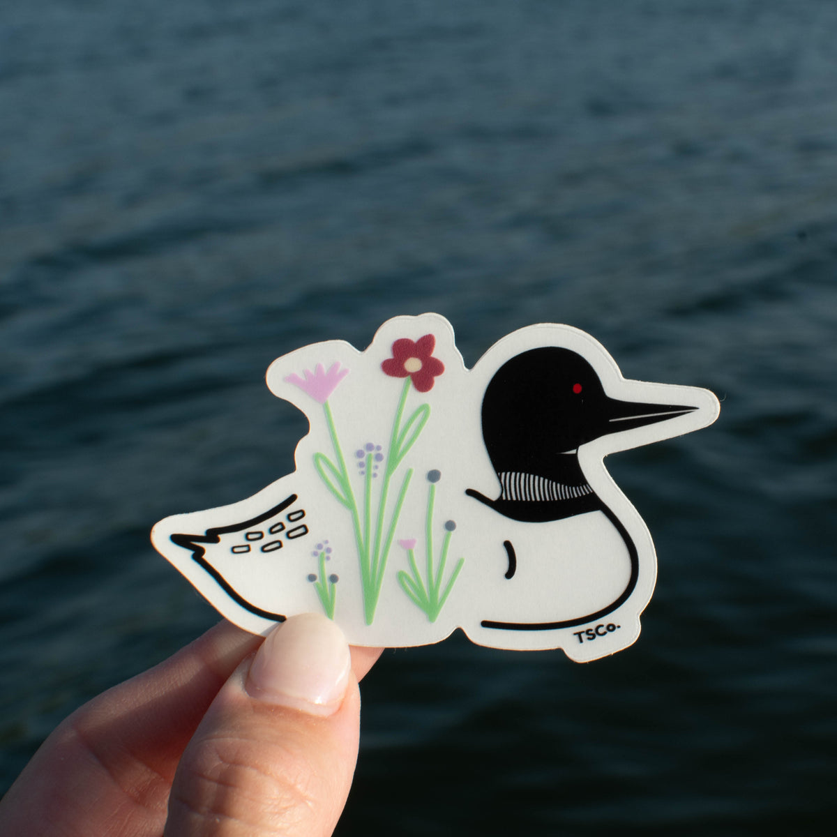 A hand holds a die-cut sticker featuring a stylized loon with flowers, set against a blurred background of dark blue water.