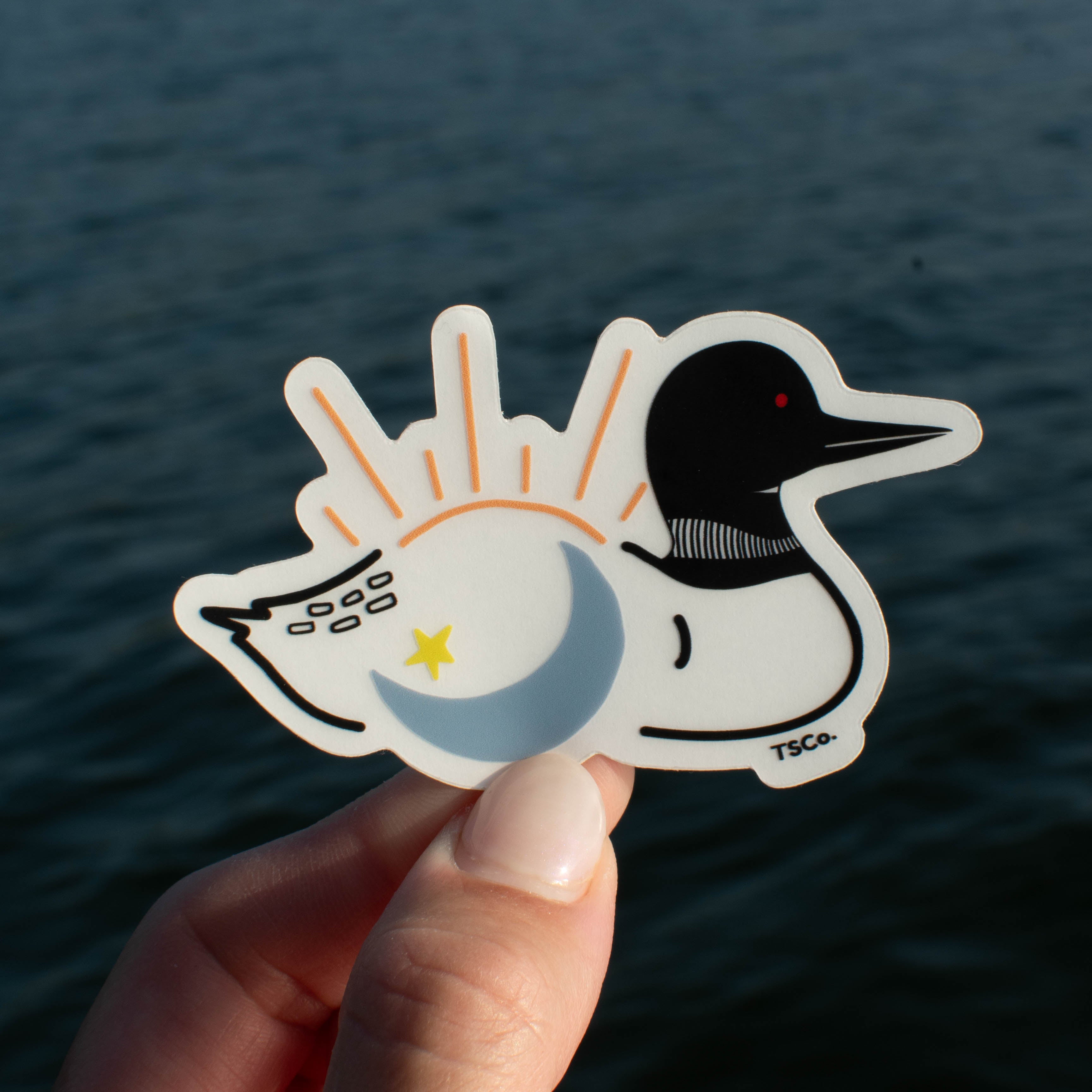 A hand holds a die-cut sticker featuring a stylized loon with a sun-and-moon motif in orange, blue, and yellow, against a blurred body of water background.
