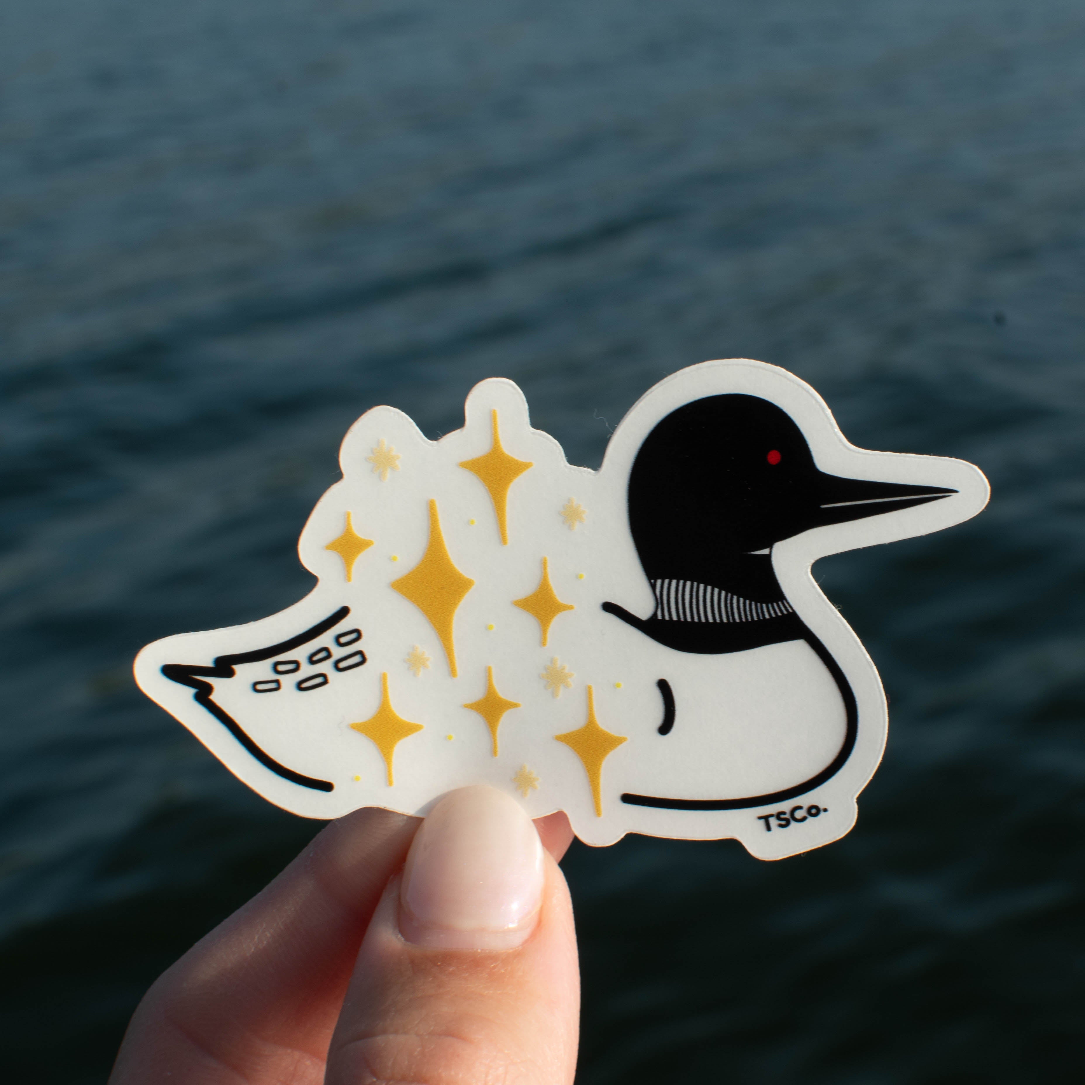 A hand holding a die-cut sticker featuring a stylized loon with gold twinkly stars on its back, set against a blurred background of dark blue water.
