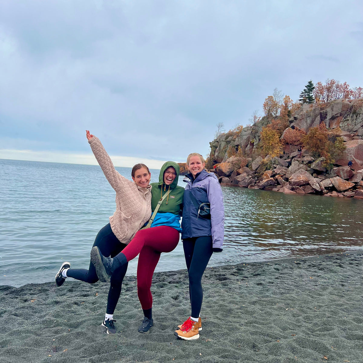 Three women posing together on a rocky beach with water and a rocky shoreline in the background, wearing colorful athletic jackets and leggings.