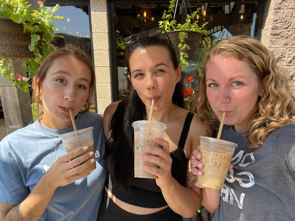 Three women are shown from a top-down angle, each holding a clear plastic cup with a straw and drinking an iced coffee in an outdoor cafe setting.