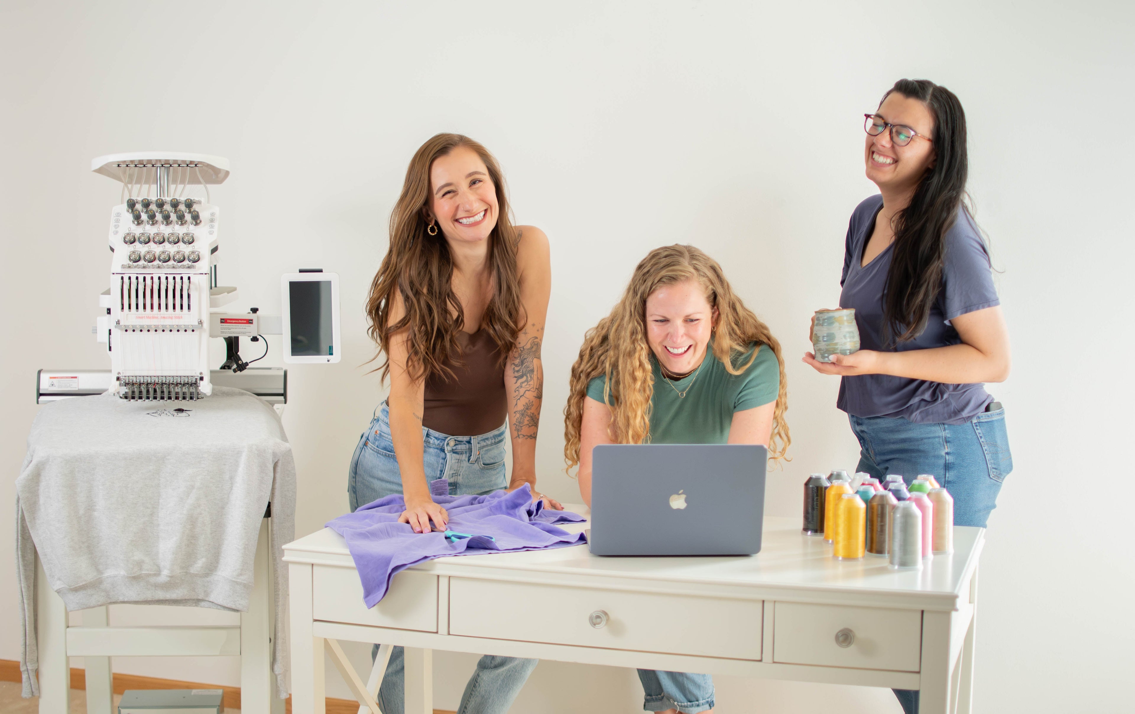 Three women in a bright studio setting with an embroidery machine, a laptop, and spools of thread. 