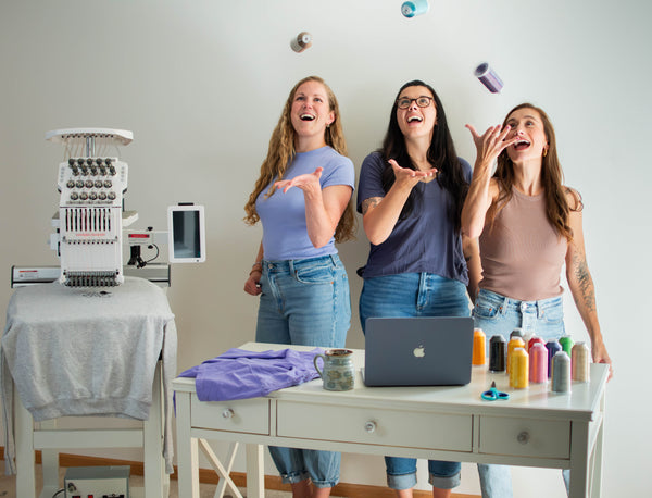Three women laughing and tossing colorful spools of thread in a bright craft room. A white embroidery machine sits on a table to the left, and a desk in the foreground holds a laptop, fabric, and multiple colored thread spools.