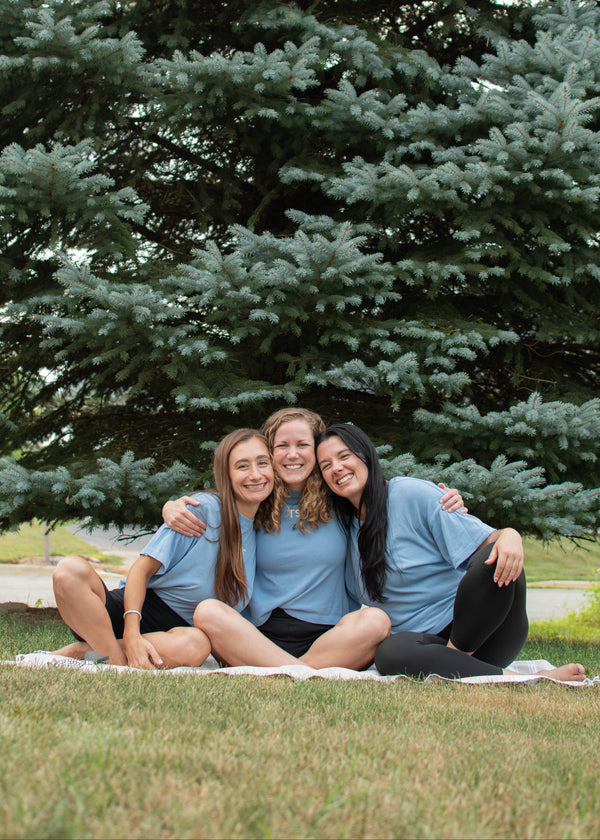 Three young women sitting together on a blanket on a grassy lawn in front of a large evergreen tree, wearing matching light blue TSCo t-shirts.