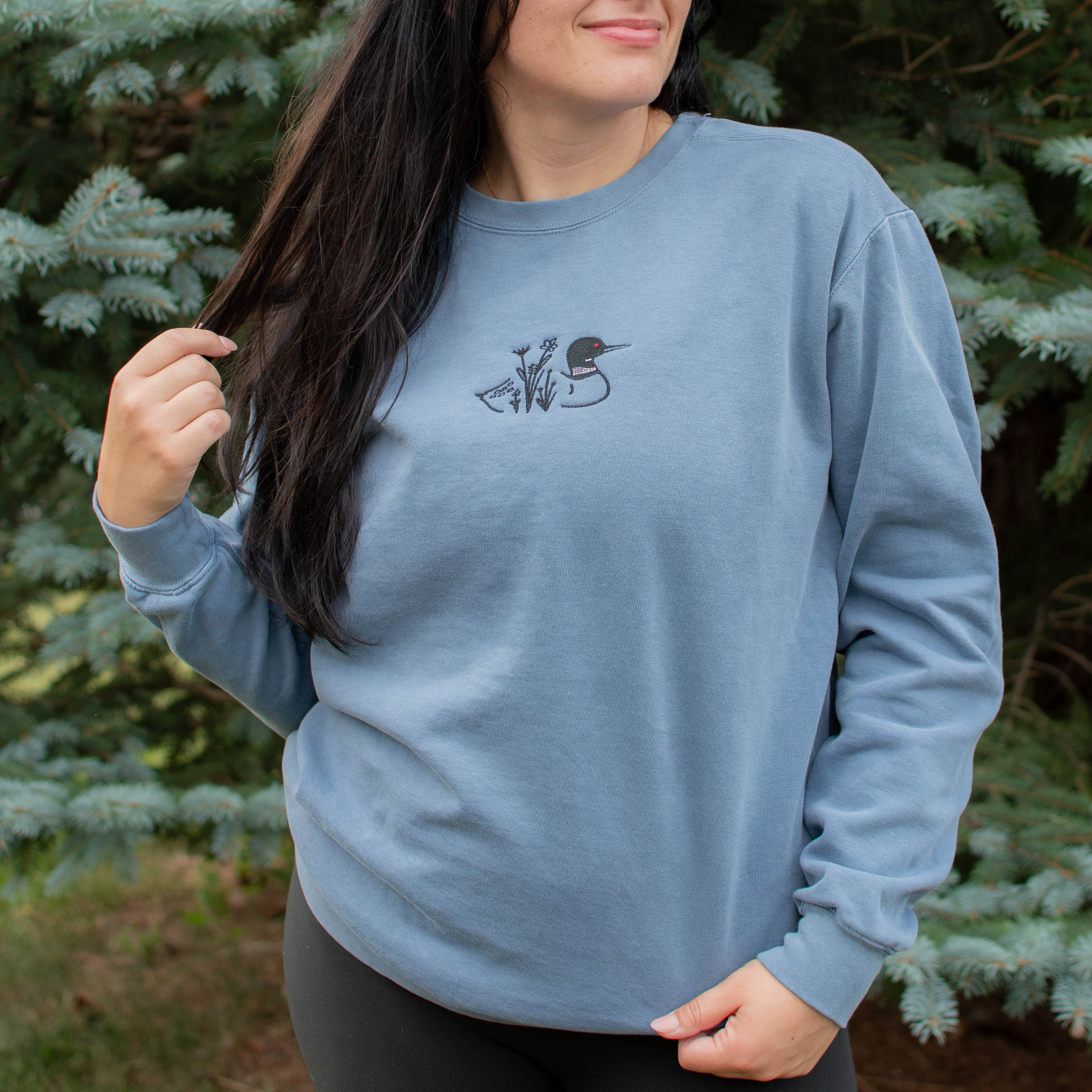 A woman wearing a blue jean colored crewneck sweatshirt with a small black embroidered loon design on the center chest, standing outdoors in front of green pine trees.