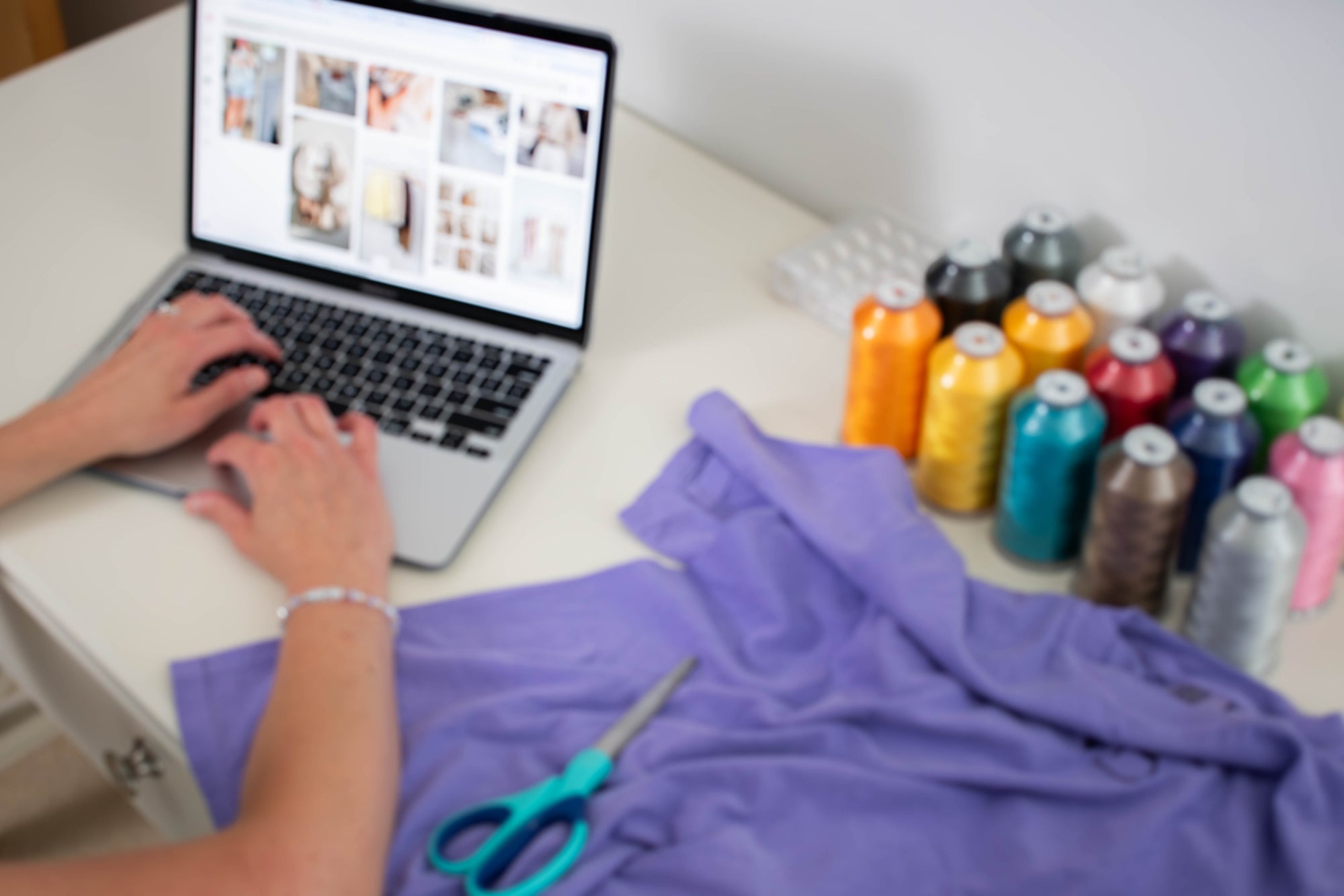 A person's hands are shown using a laptop on a white desk, with a purple fabric piece in the foreground, teal scissors, and multiple colorful spools of thread arranged on the right side.