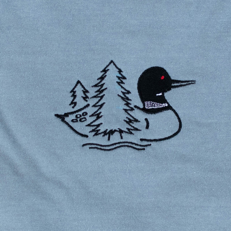 A close-up of a warm gray fabric surface featuring a black embroidered loon design with two evergreen trees and wavy water lines with a small red eye detail.