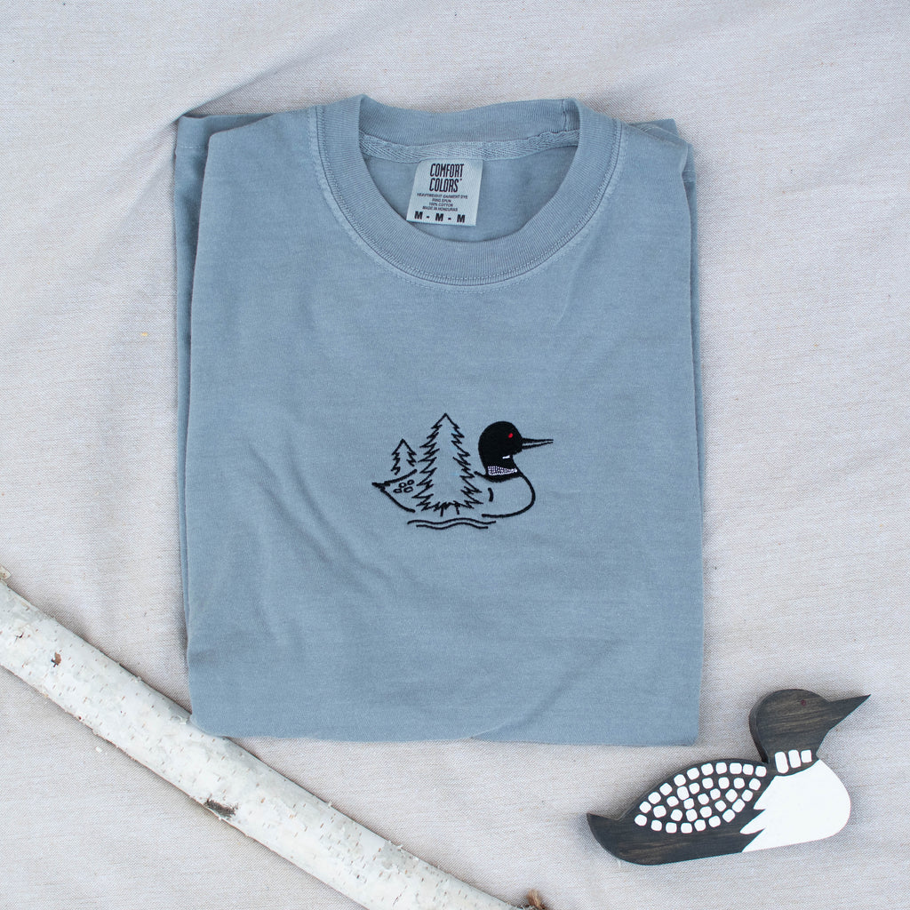 A folded granite colored sweatshirt with with a small black embroidered loon design on the center chest, displayed on a light tan fabric background with a decorative wooden loon and a white birch branch.