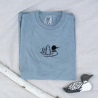A folded granite colored sweatshirt with with a small black embroidered loon design on the center chest, displayed on a light tan fabric background with a decorative wooden loon and a white birch branch.