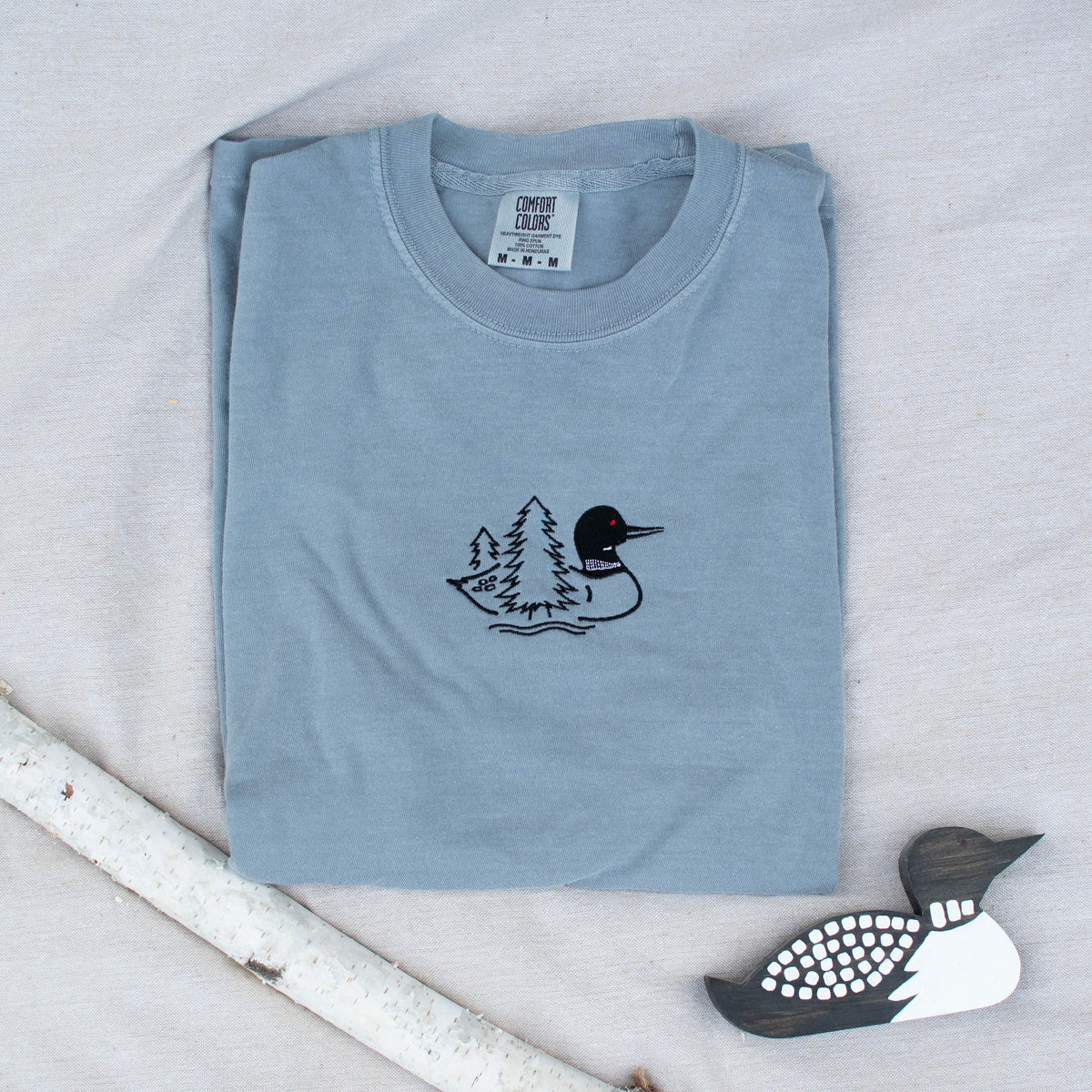 A folded granite colored sweatshirt with with a small black embroidered loon design on the center chest, displayed on a light tan fabric background with a decorative wooden loon and a white birch branch.