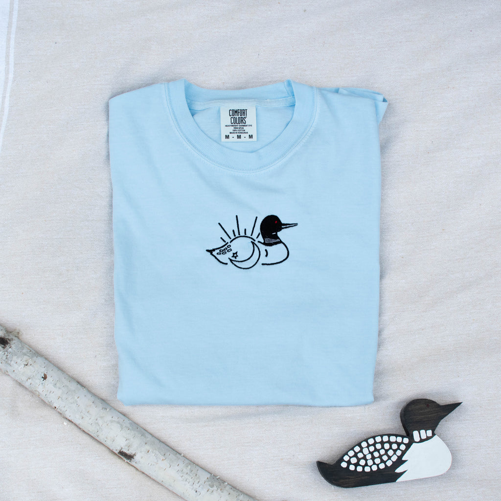 A folded chambray colored sweatshirt with with a small black embroidered loon design on the center chest, displayed on a light tan fabric background with a decorative wooden loon and a white birch branch.