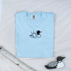A folded chambray colored sweatshirt with with a small black embroidered loon design on the center chest, displayed on a light tan fabric background with a decorative wooden loon and a white birch branch.