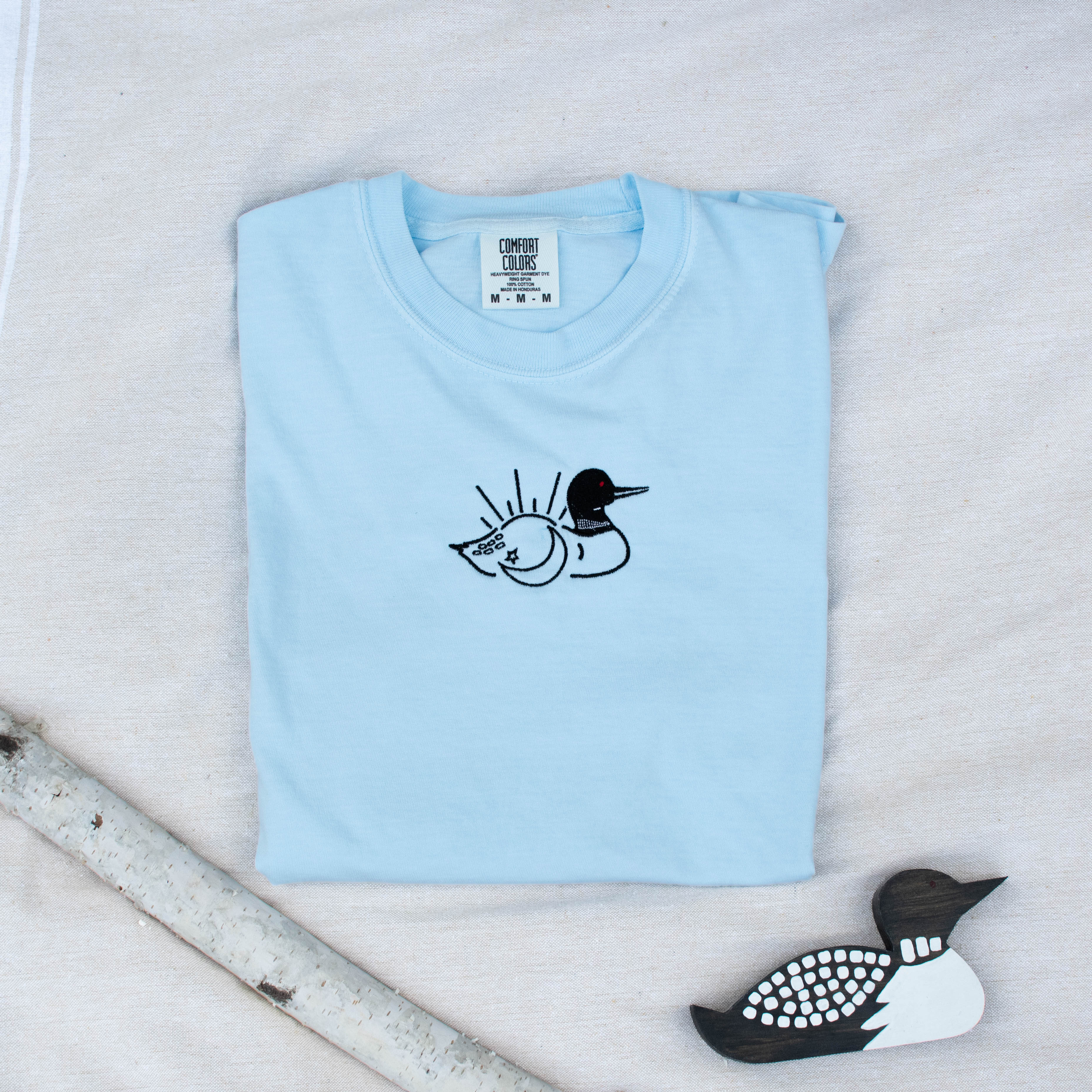 A folded chambray colored sweatshirt with with a small black embroidered loon design on the center chest, displayed on a light tan fabric background with a decorative wooden loon and a white birch branch.