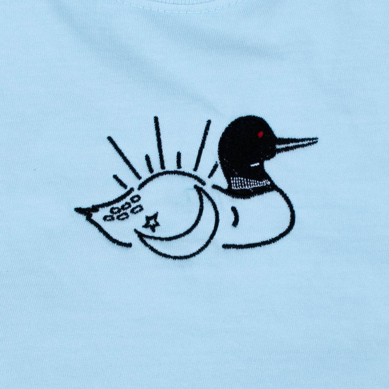A close-up of a light blue fabric surface featuring a black embroidered loon design with a crescent moon, star, and sunset radiating lines behind it with a small red eye detail.