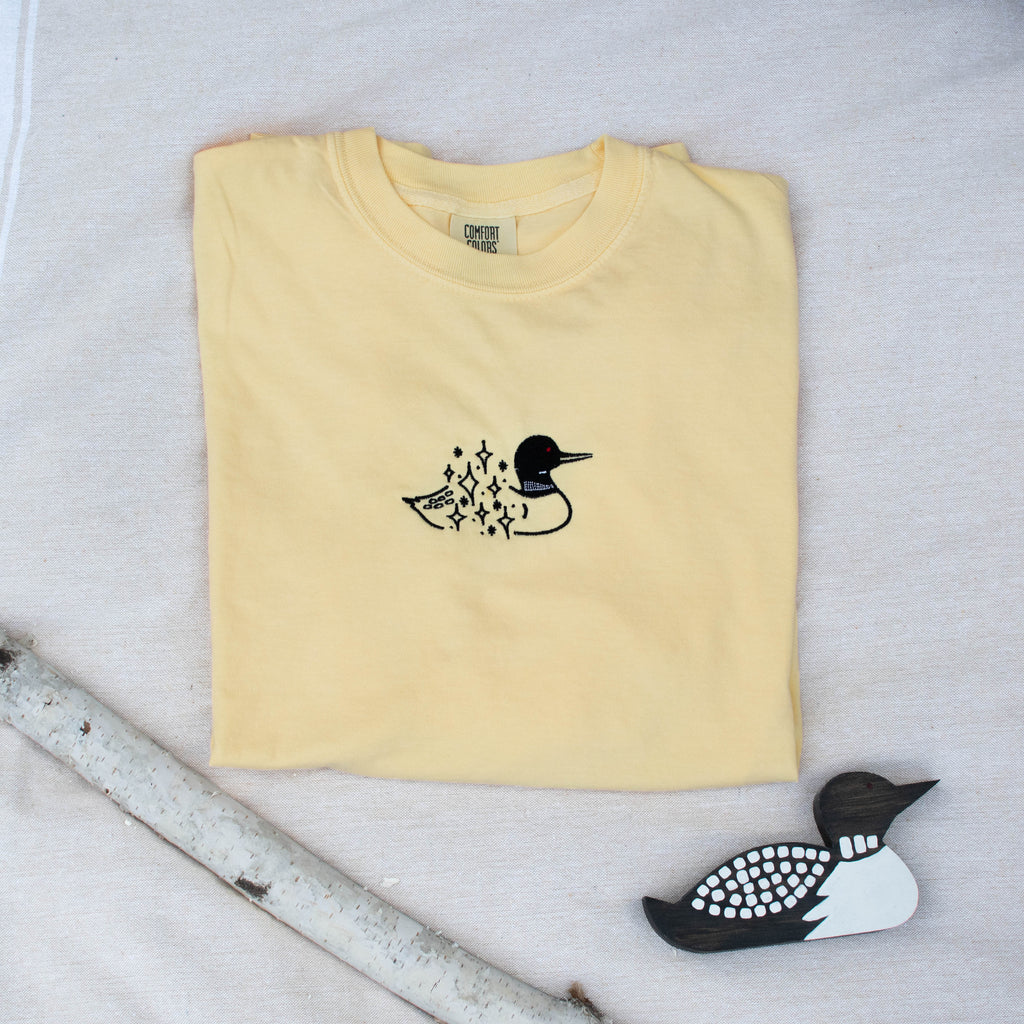 A folded butter colored sweatshirt with with a small black embroidered loon design on the center chest, displayed on a light tan fabric background with a decorative wooden loon and a white birch branch.