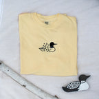 A folded butter colored sweatshirt with with a small black embroidered loon design on the center chest, displayed on a light tan fabric background with a decorative wooden loon and a white birch branch.