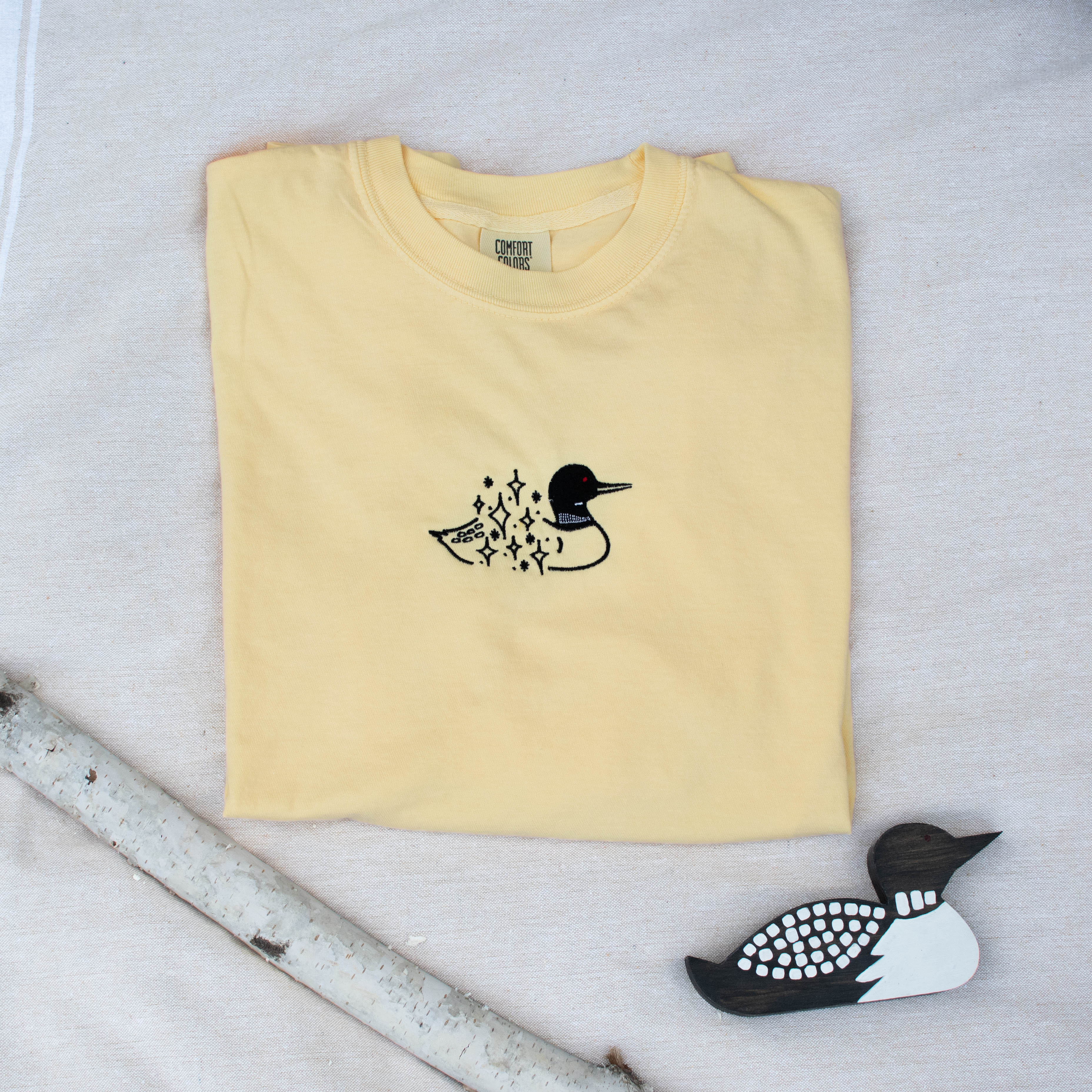 A folded butter colored sweatshirt with with a small black embroidered loon design on the center chest, displayed on a light tan fabric background with a decorative wooden loon and a white birch branch.