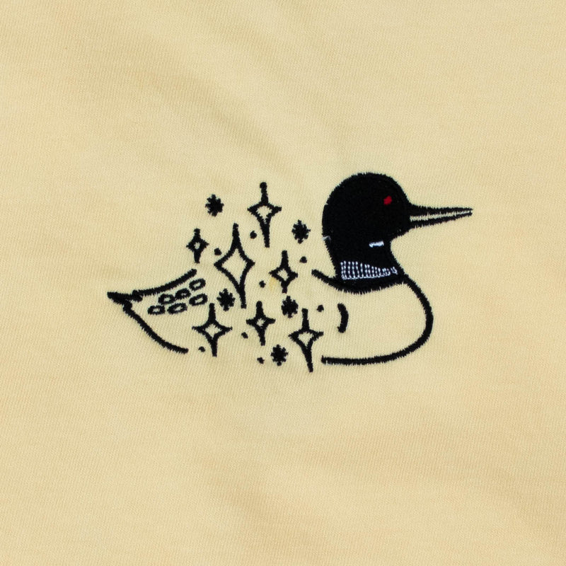 A close-up of a light yellow fabric surface featuring a black embroidered loon design with twinkly stars on the body with a small red eye detail.