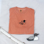 A folded yam colored sweatshirt with with a small black embroidered loon design on the center chest, displayed on a light tan fabric background with a decorative wooden loon and a white birch branch.