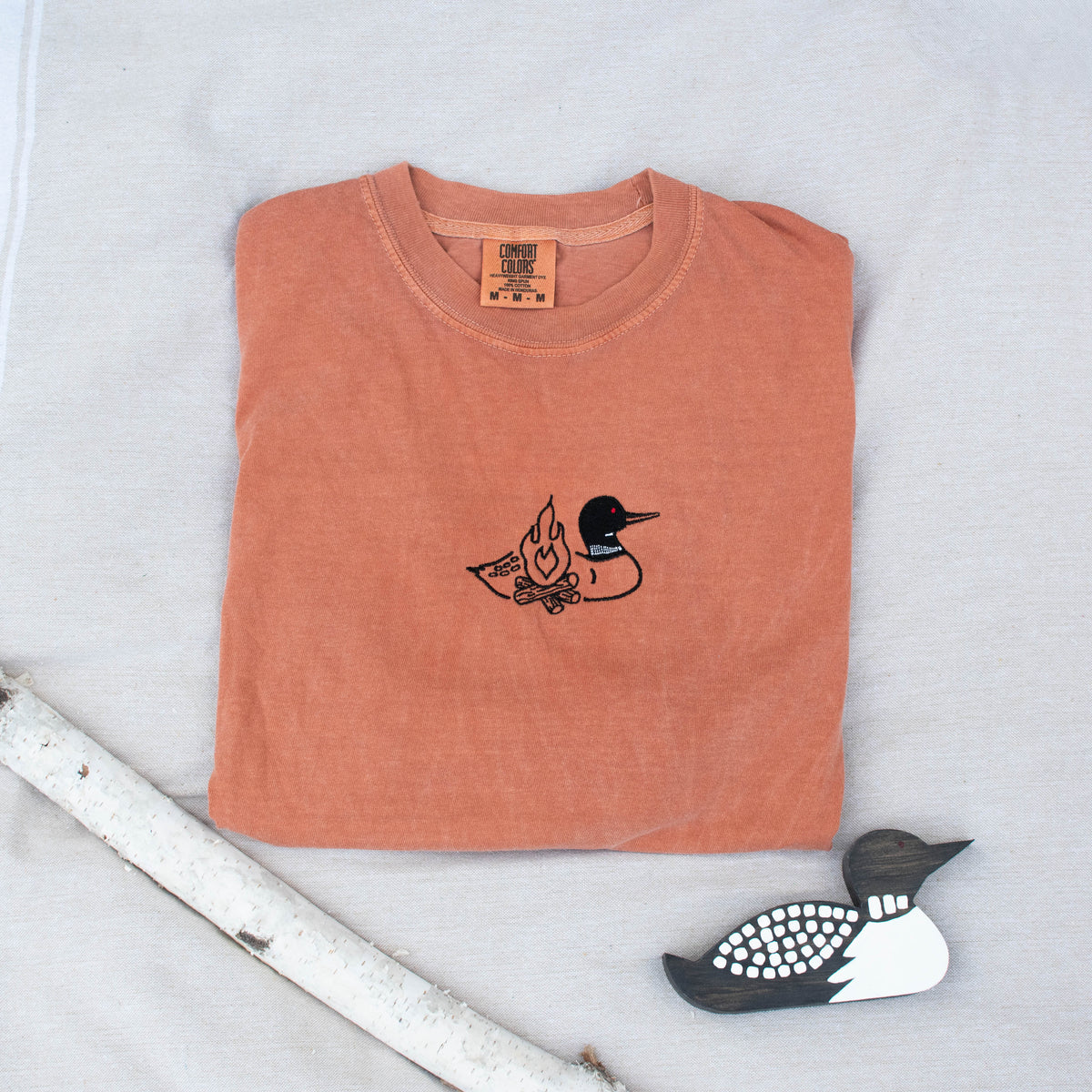 A folded yam colored sweatshirt with with a small black embroidered loon design on the center chest, displayed on a light tan fabric background with a decorative wooden loon and a white birch branch.