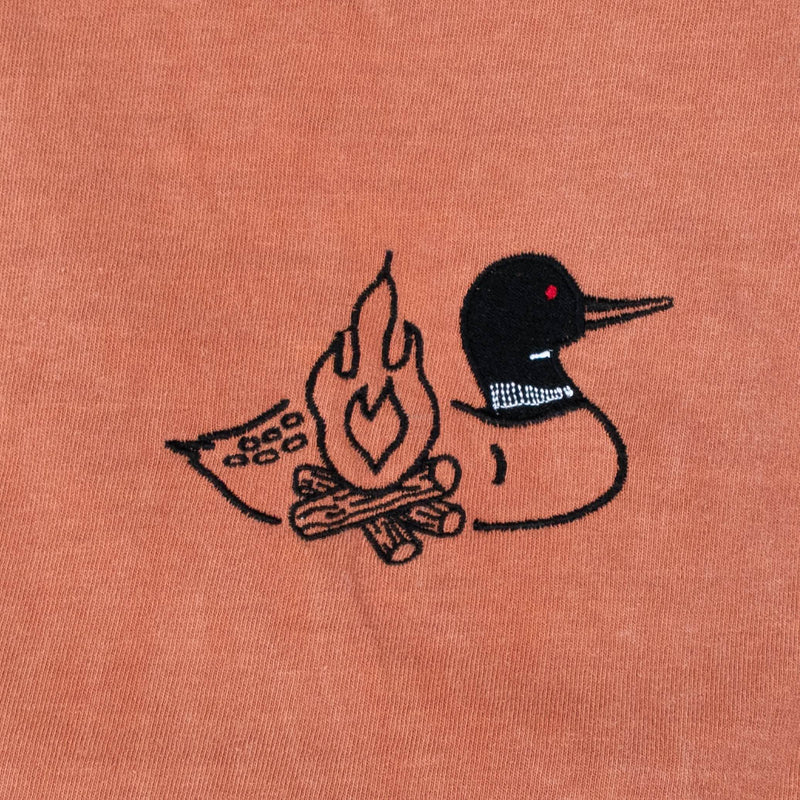 A close-up of a terracotta-colored fabric surface featuring a black embroidered loon design with a campfire and logs on its body with a small red eye detail.