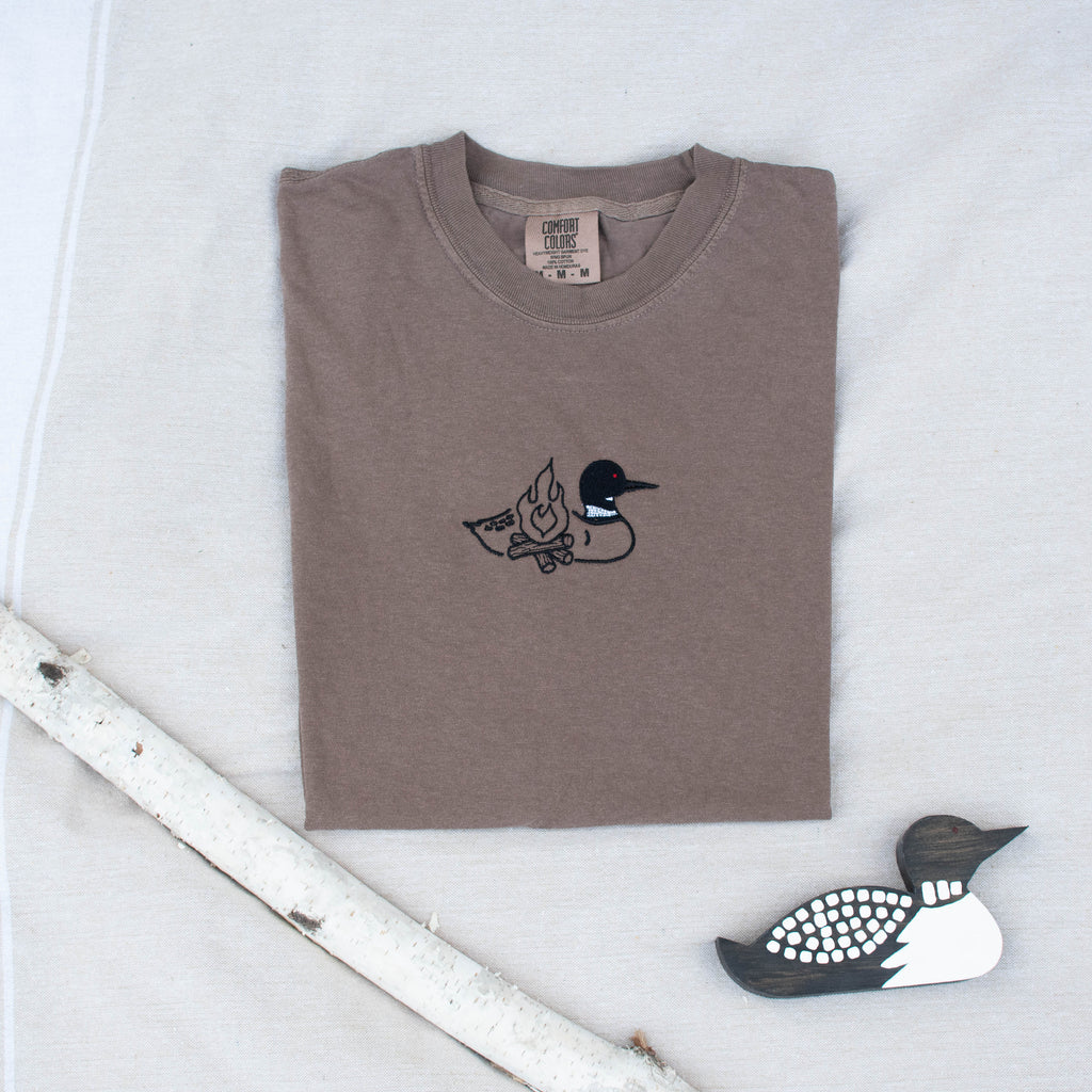 A folded espresso colored sweatshirt with with a small black embroidered loon design on the center chest, displayed on a light tan fabric background with a decorative wooden loon and a white birch branch.