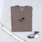 A folded espresso colored sweatshirt with with a small black embroidered loon design on the center chest, displayed on a light tan fabric background with a decorative wooden loon and a white birch branch.