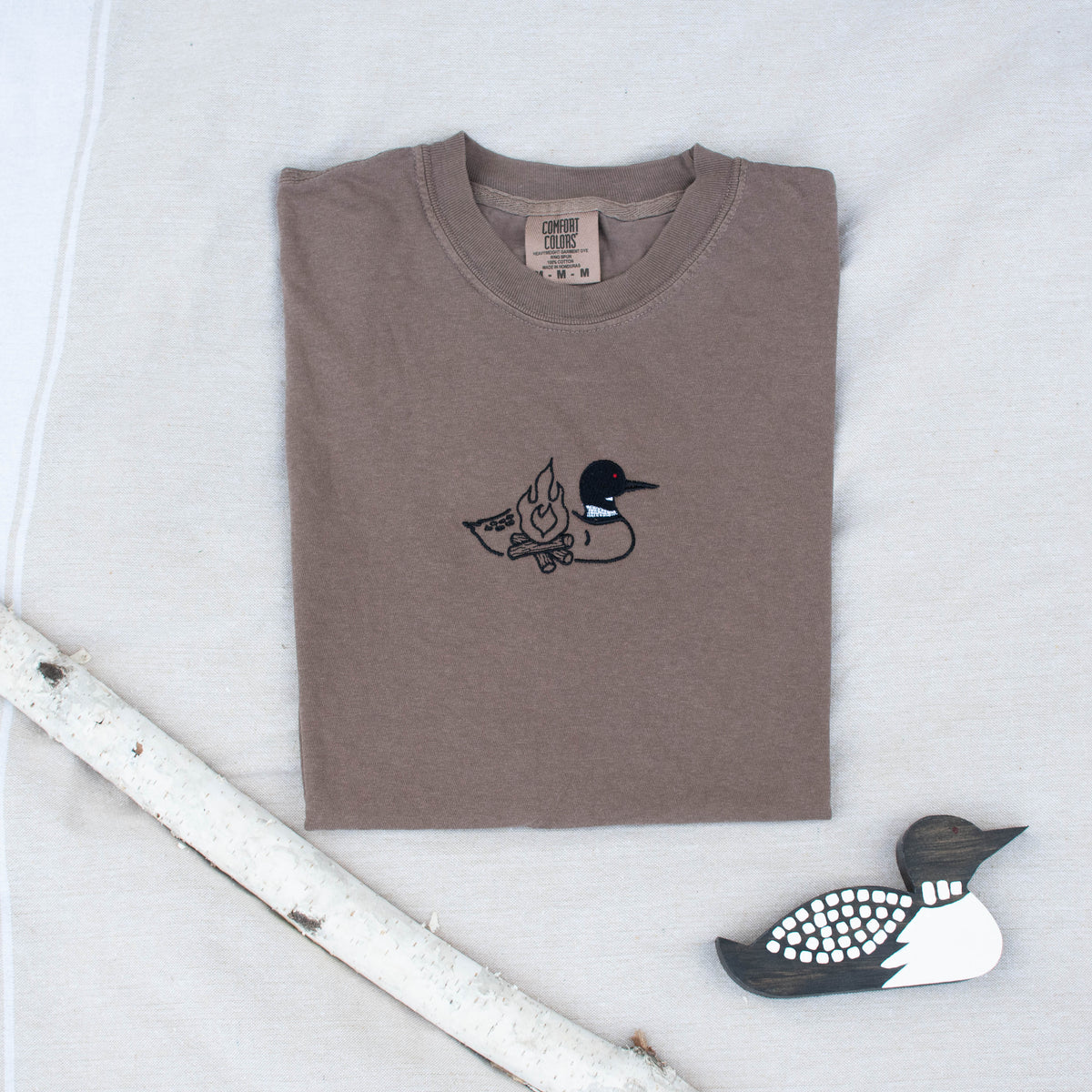 A folded espresso colored sweatshirt with with a small black embroidered loon design on the center chest, displayed on a light tan fabric background with a decorative wooden loon and a white birch branch.