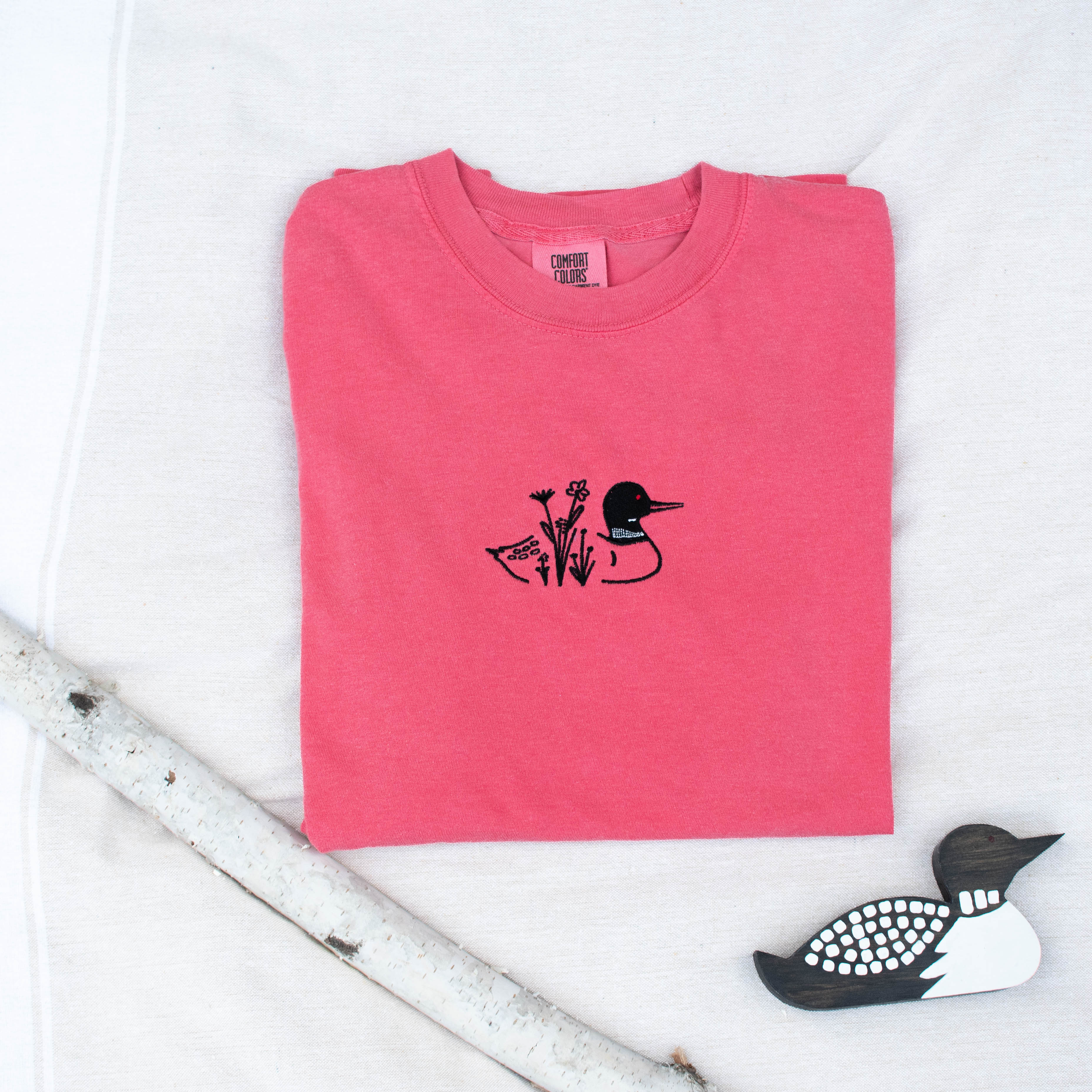 A folded watermelon colored sweatshirt with with a small black embroidered loon design on the center chest, displayed on a light tan fabric background with a decorative wooden loon and a white birch branch.