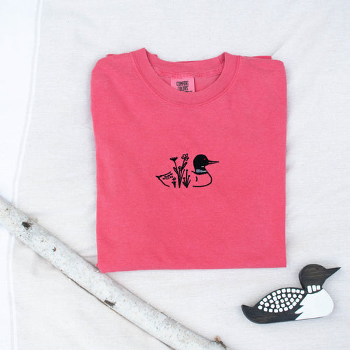 A folded watermelon colored sweatshirt with with a small black embroidered loon design on the center chest, displayed on a light tan fabric background with a decorative wooden loon and a white birch branch.