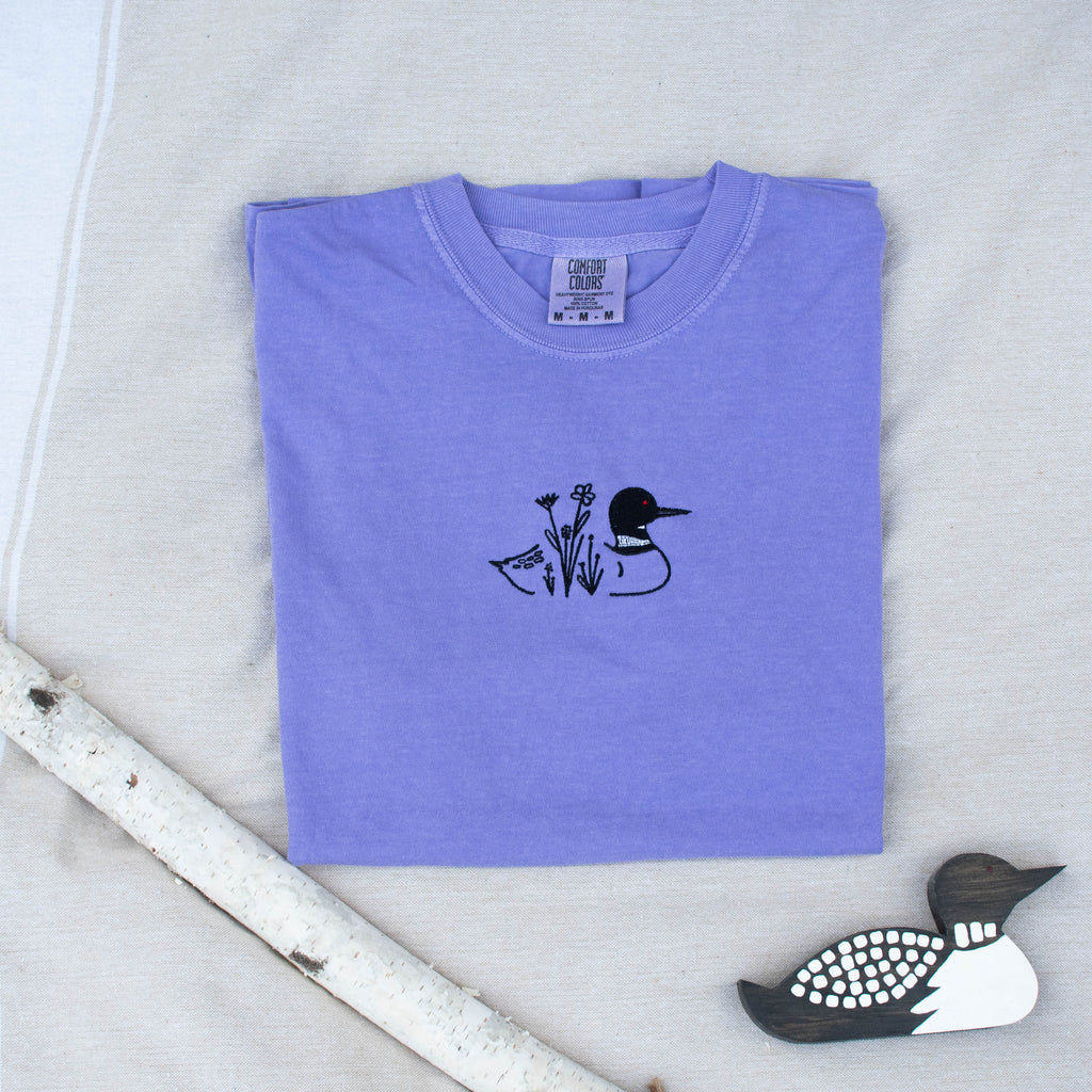 A folded violet colored sweatshirt with with a small black embroidered loon design on the center chest, displayed on a light tan fabric background with a decorative wooden loon and a white birch branch.