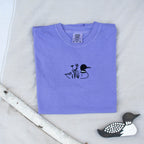 A folded violet colored sweatshirt with with a small black embroidered loon design on the center chest, displayed on a light tan fabric background with a decorative wooden loon and a white birch branch.