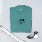 A folded light green colored sweatshirt with with a small black embroidered loon design on the center chest, displayed on a light tan fabric background with a decorative wooden loon and a white birch branch.
