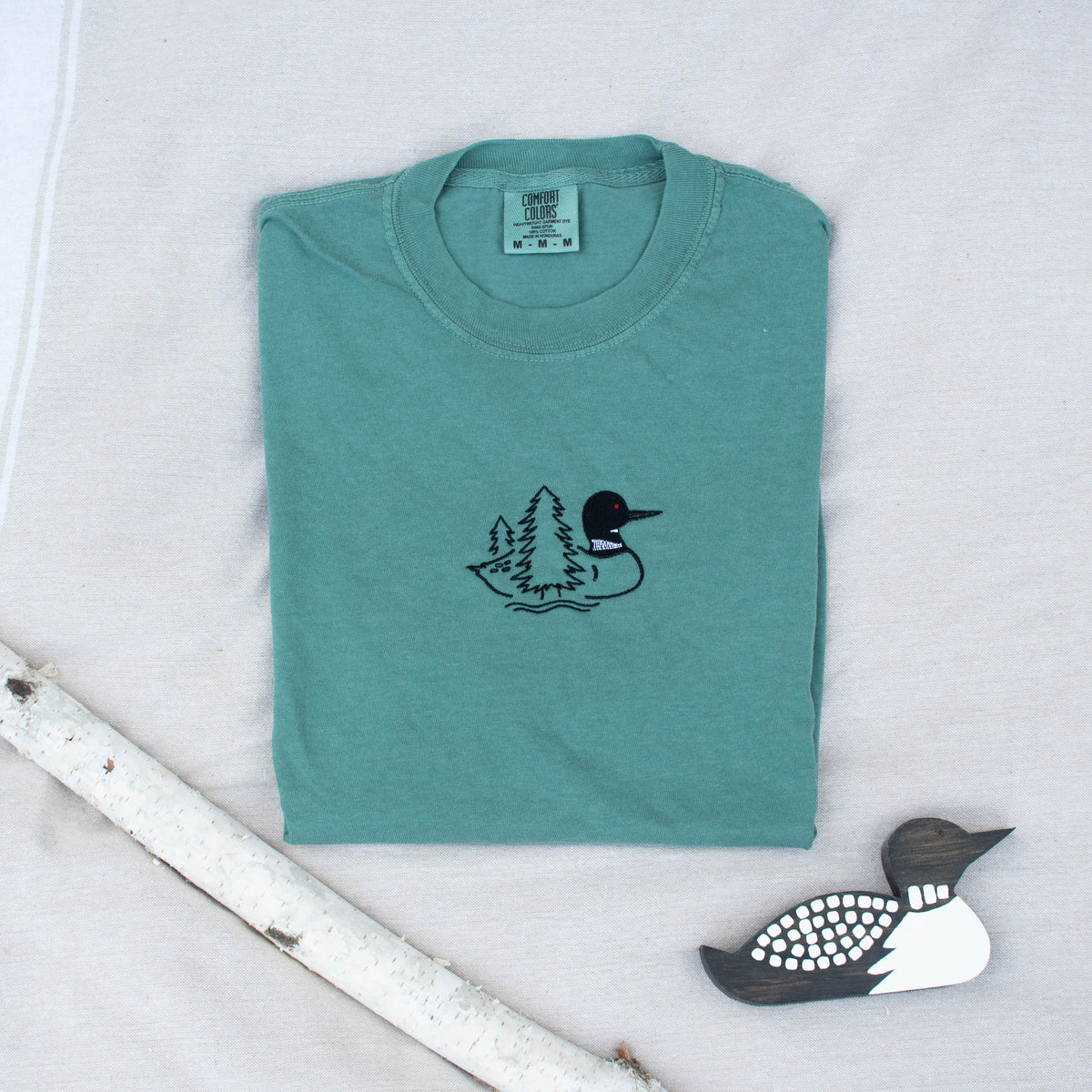 A folded light green colored sweatshirt with with a small black embroidered loon design on the center chest, displayed on a light tan fabric background with a decorative wooden loon and a white birch branch.