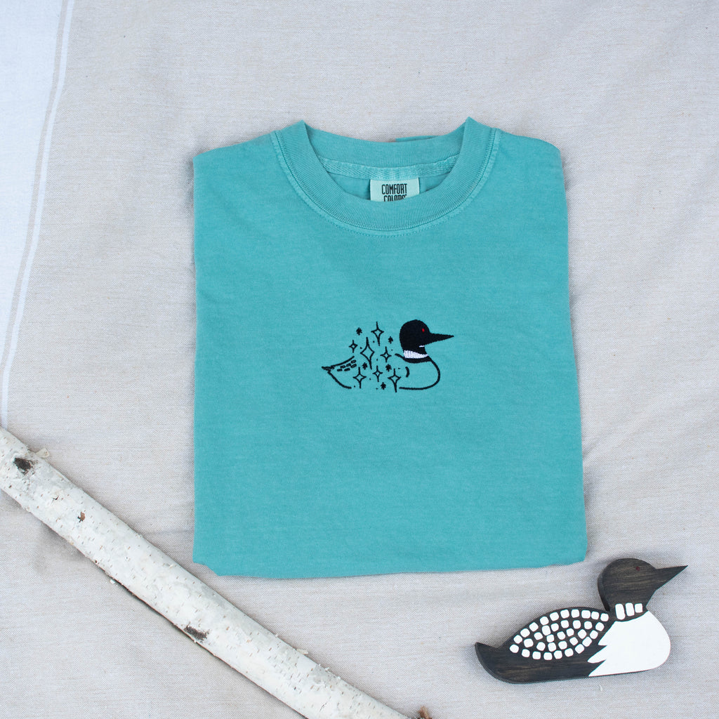 A folded seafoam colored sweatshirt with with a small black embroidered loon design on the center chest, displayed on a light tan fabric background with a decorative wooden loon and a white birch branch.