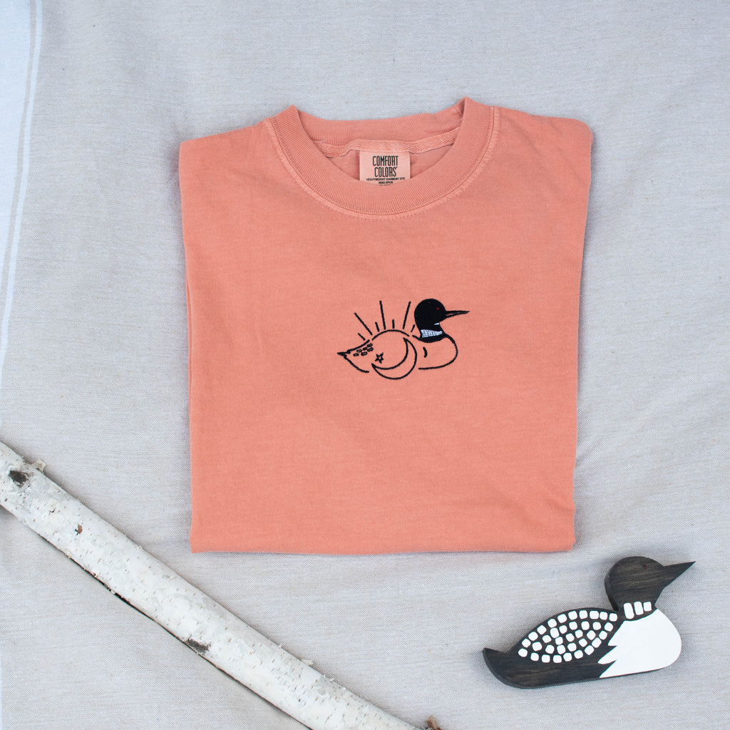 A folded terracotta colored sweatshirt with with a small black embroidered loon design on the center chest, displayed on a light tan fabric background with a decorative wooden loon and a white birch branch.
