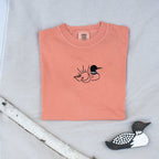 A folded terracotta colored sweatshirt with with a small black embroidered loon design on the center chest, displayed on a light tan fabric background with a decorative wooden loon and a white birch branch.