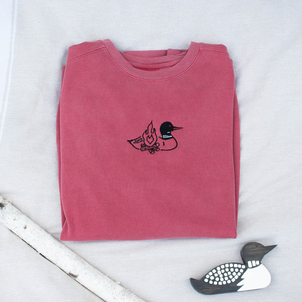 A folded crimson colored sweatshirt with with a small black embroidered loon design on the center chest, displayed on a light tan fabric background with a decorative wooden loon and a white birch branch.