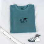 A folded blue spruce colored sweatshirt with with a small black embroidered loon design on the center chest, displayed on a light tan fabric background with a decorative wooden loon and a white birch branch.