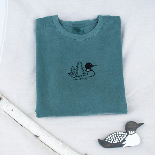 A folded blue spruce colored sweatshirt with with a small black embroidered loon design on the center chest, displayed on a light tan fabric background with a decorative wooden loon and a white birch branch.