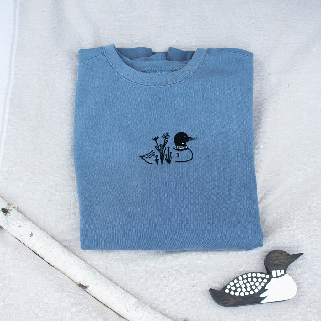 A folded blue jean colored sweatshirt with with a small black embroidered loon design on the center chest, displayed on a light tan fabric background with a decorative wooden loon and a white birch branch.