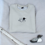 A folded sand colored sweatshirt with with a small black embroidered loon design on the center chest, displayed on a light tan fabric background with a decorative wooden loon and a white birch branch.