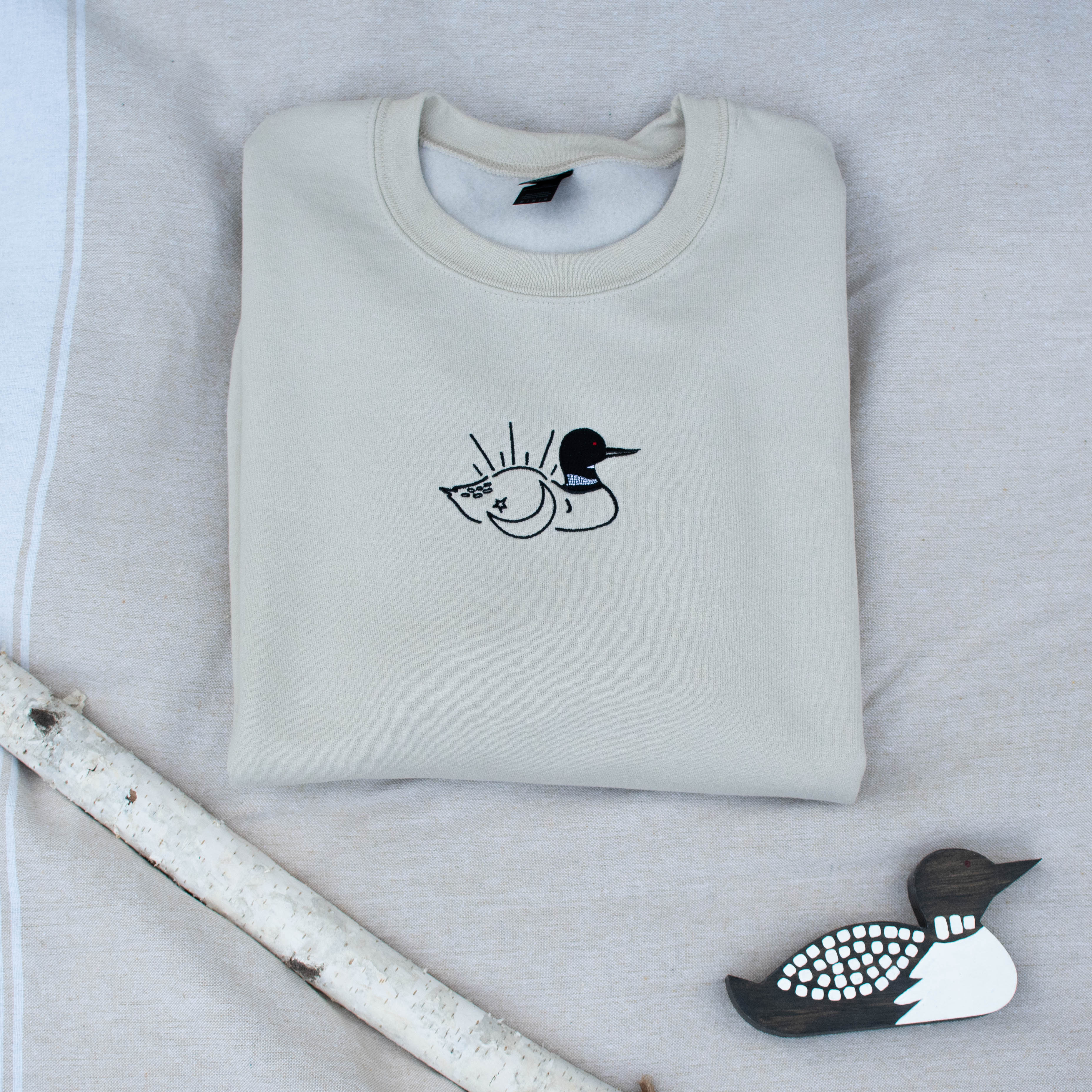 A folded sand colored sweatshirt with with a small black embroidered loon design on the center chest, displayed on a light tan fabric background with a decorative wooden loon and a white birch branch.
