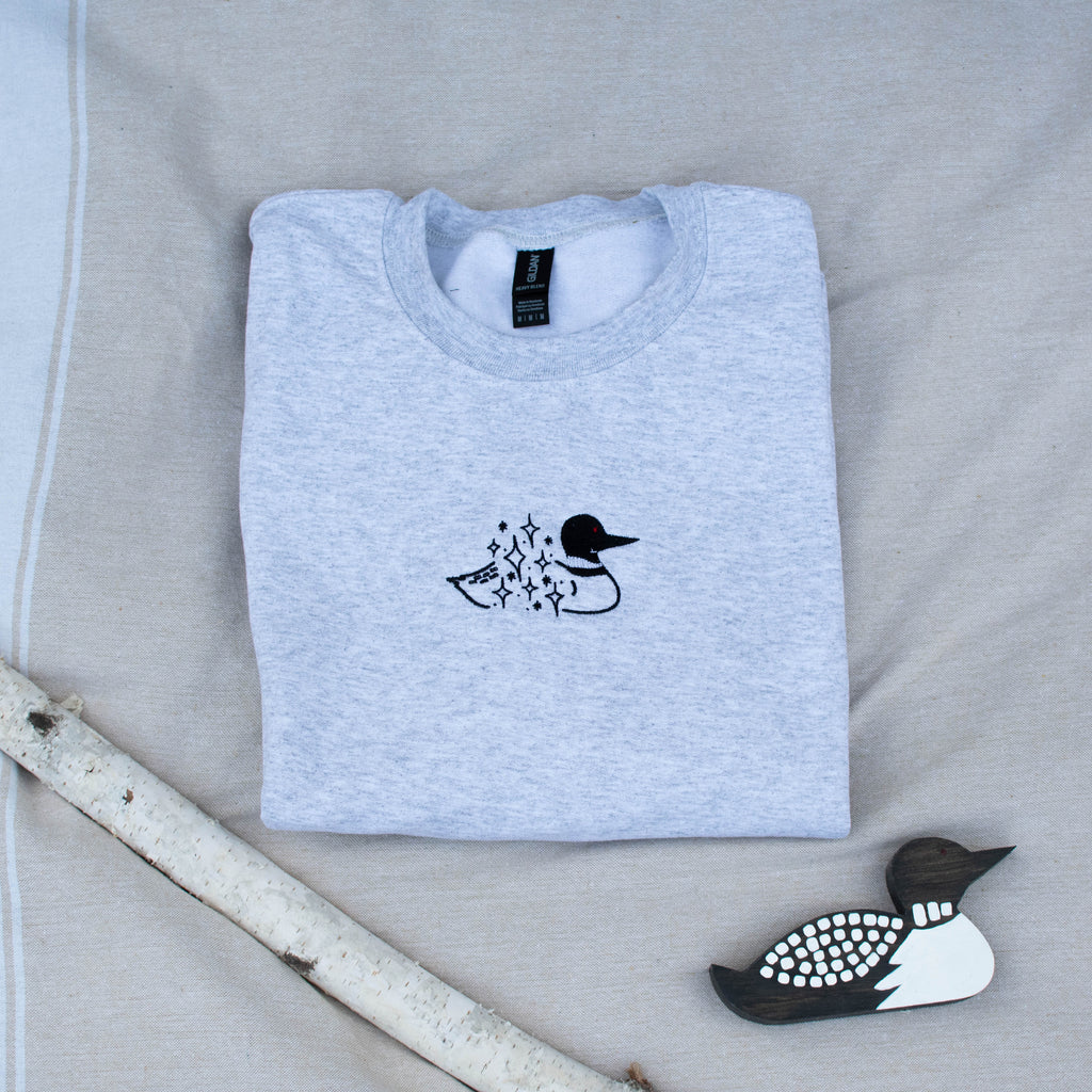 A folded ash colored sweatshirt with with a small black embroidered loon design on the center chest, displayed on a light tan fabric background with a decorative wooden loon and a white birch branch.