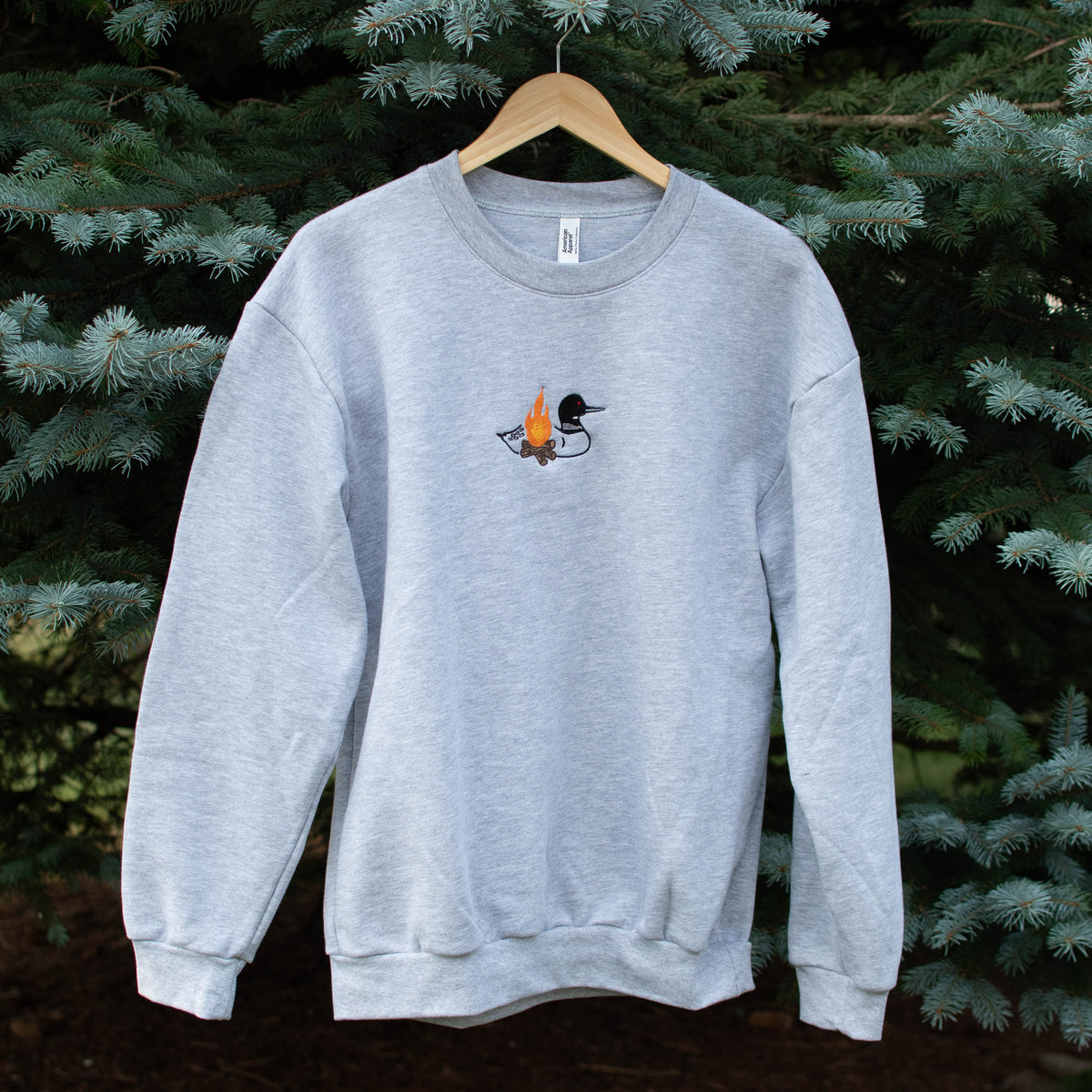 A light heather gray crewneck sweatshirt hanging on a wooden hanger against a background of green pine trees. The sweatshirt features a small embroidered loon design on the center chest.