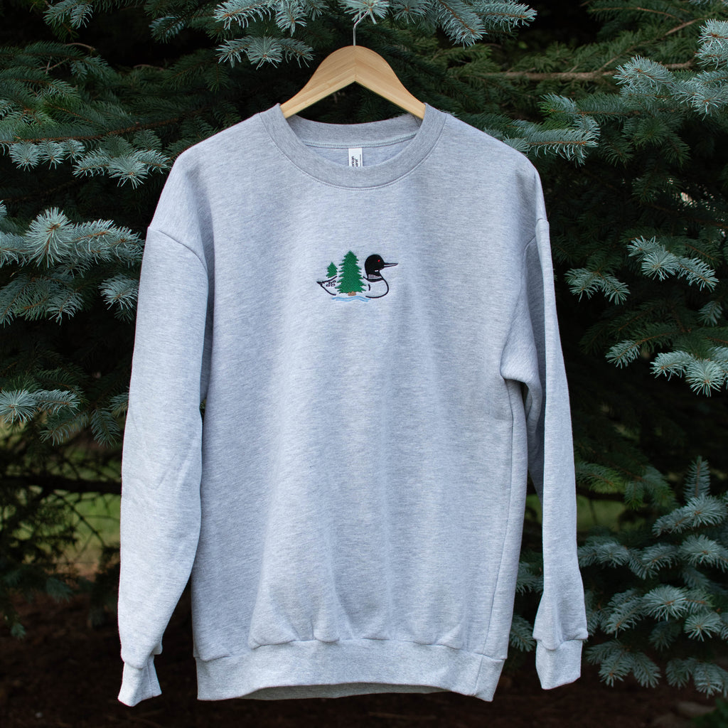 A light heather gray crewneck sweatshirt hanging on a wooden hanger against a background of green pine trees. The sweatshirt features a small embroidered loon design on the center chest.