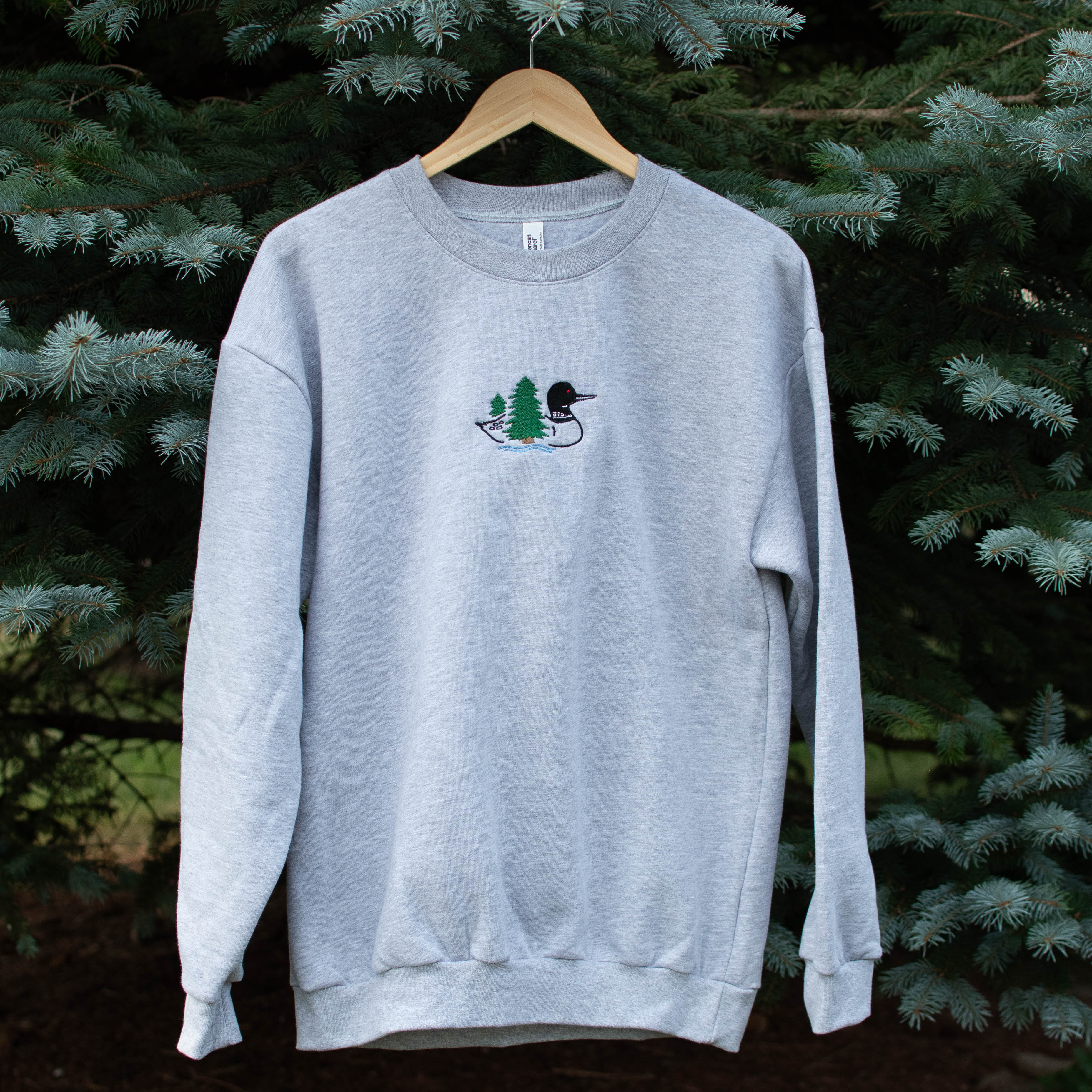 A light heather gray crewneck sweatshirt hanging on a wooden hanger against a background of green pine trees. The sweatshirt features a small embroidered loon design on the center chest.