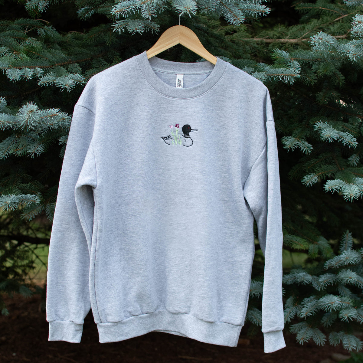 A light heather gray crewneck sweatshirt hanging on a wooden hanger against a background of green pine trees. The sweatshirt features a small embroidered loon design on the center chest.