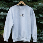 A light heather gray crewneck sweatshirt hanging on a wooden hanger against a background of green pine trees. The sweatshirt features a small embroidered loon design on the center chest.